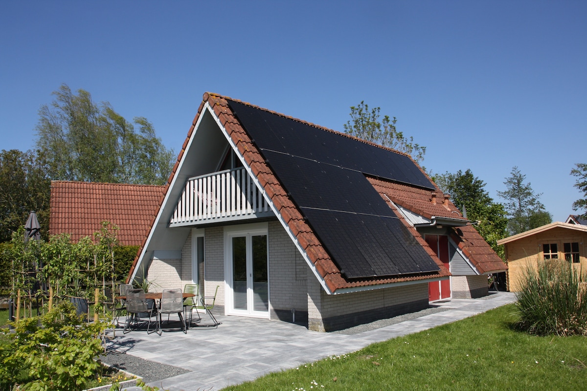 The exterior of a bungalow features a sloped roof adorned with solar panels. A spacious patio area with outdoor seating is visible, surrounded by greenery. The building is constructed with light-colored materials and has large windows that allow for natural light.