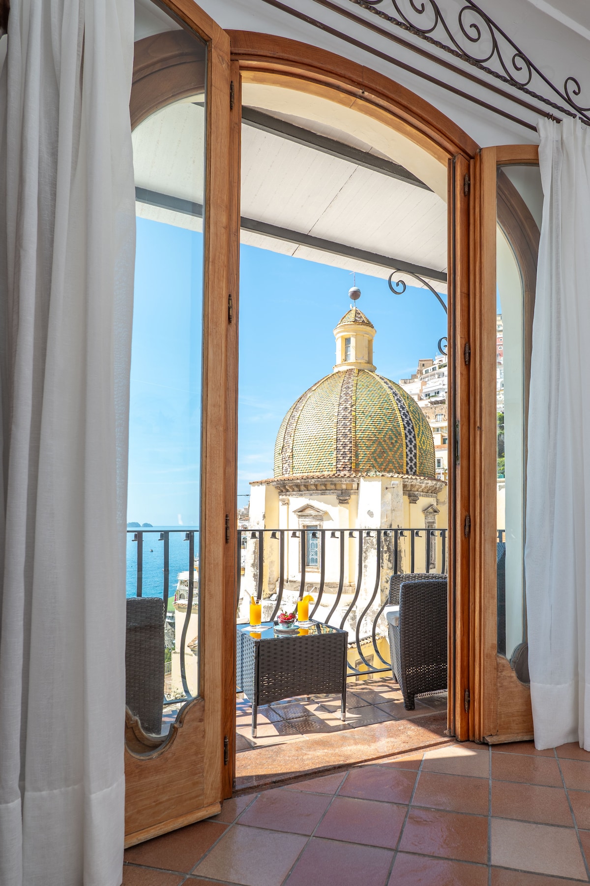 A doorway frames a scenic view of a domed building adorned with intricate tiles, contrasting against a clear blue sky. Outdoor seating is positioned on a balcony, complemented by glasses, suggesting a peaceful space for relaxation.