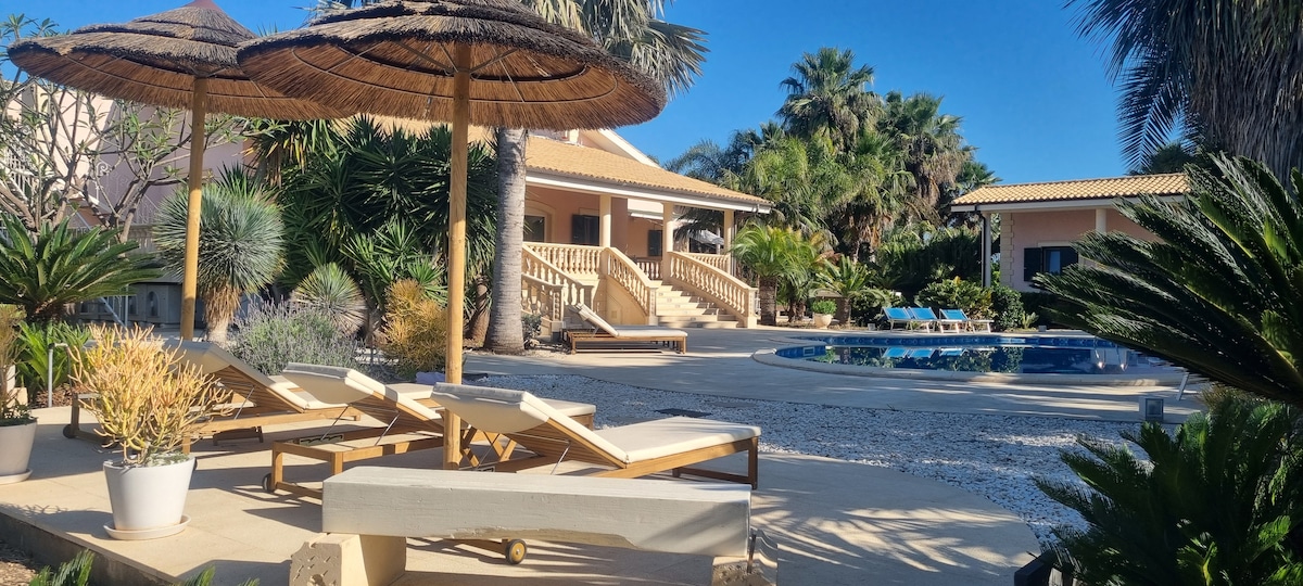 A spacious outdoor area features loungers and umbrellas beside a swimming pool, surrounded by tropical plants. The setting includes a sun-drenched terrace, with a two-story building in the background. Pathways are lined with greenery, contributing to a serene environment.