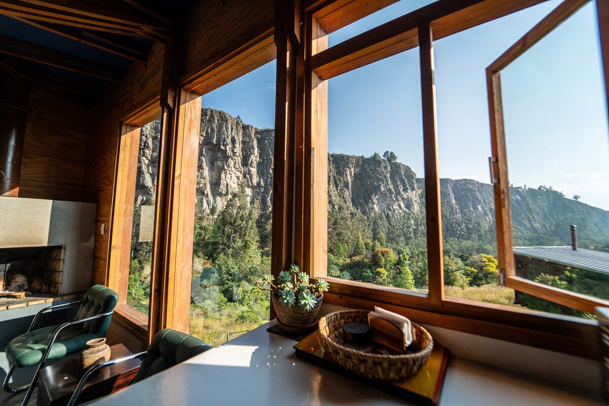 Large windows frame a breathtaking view of the rocky cliffs and lush greenery outside. Natural light fills the space, highlighting wooden accents and a cozy seating area with chairs. A simple table is set with a small decorative tray, adding to the inviting atmosphere.