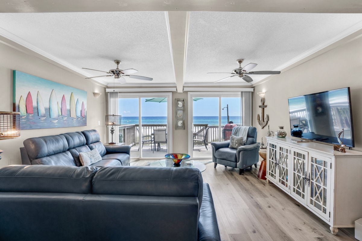 The spacious living area features comfortable seating and large windows offering ocean views. A colorful wall art piece depicting sailboats adds a touch of character. Light streams in, enhancing the airy atmosphere, while a modern television and decorative furnishings complete the inviting space.