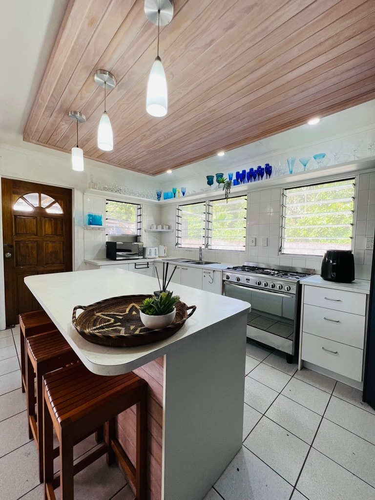 A modern kitchen features white cabinetry and a central island with bar stools. Brightly colored glassware adorns the shelves, while natural light filters through the windowed walls. Stainless steel appliances, including a stove and microwave, provide functionality in the space.