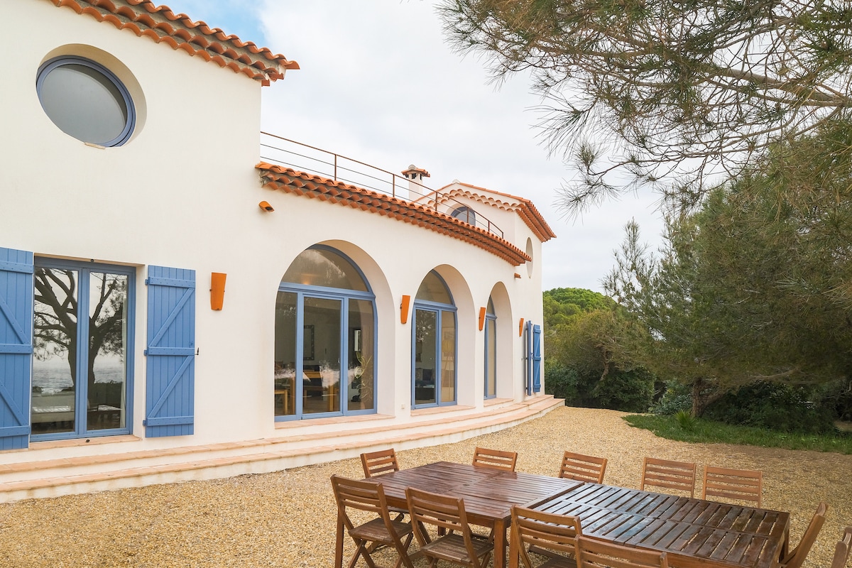 The exterior of the villa features a white façade with arched windows and blue shutters. A spacious outdoor area includes a large wooden dining table surrounded by matching chairs, ideal for enjoying meals al fresco. Lush greenery and pine trees provide a serene backdrop.