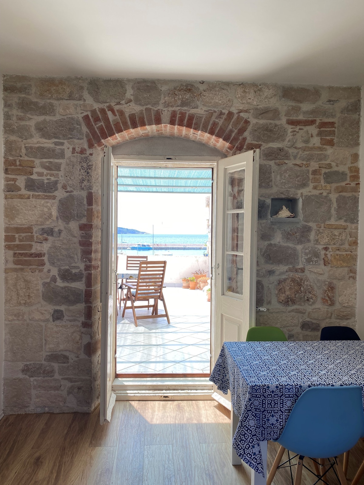 An arched doorway with white-framed doors opens to a terrace, revealing views of the sea. Stone walls contrast with a patterned tablecloth on a dining table and colorful chairs. Sunlight illuminates the entryway, creating a bright transition between the indoor and outdoor spaces.