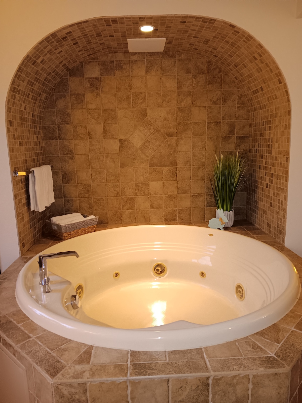 A spacious jacuzzi tub is set against a beautifully tiled wall, with natural tones enhancing the calming ambiance. Towels are neatly arranged on a nearby holder, while a small plant adds a touch of greenery to the serene space.