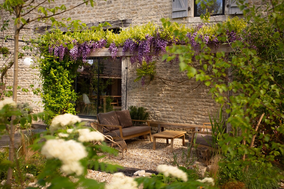 A serene outdoor seating area is framed by lush greenery and vibrant purple wisteria. Comfortable chairs surround a wooden table, offering a relaxing space within a gravel courtyard. The stone wall of the house provides a rustic backdrop, enhancing the tranquil garden setting.