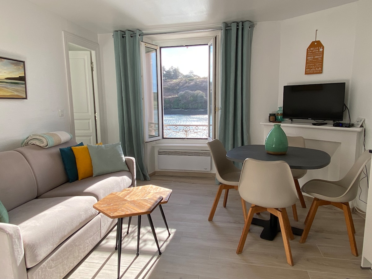 A bright living area features a light-colored sofa adorned with decorative pillows. A small wooden coffee table sits in front of a round dining table with four chairs. Large windows frame a scenic view of the port, with gentle light illuminating the space.