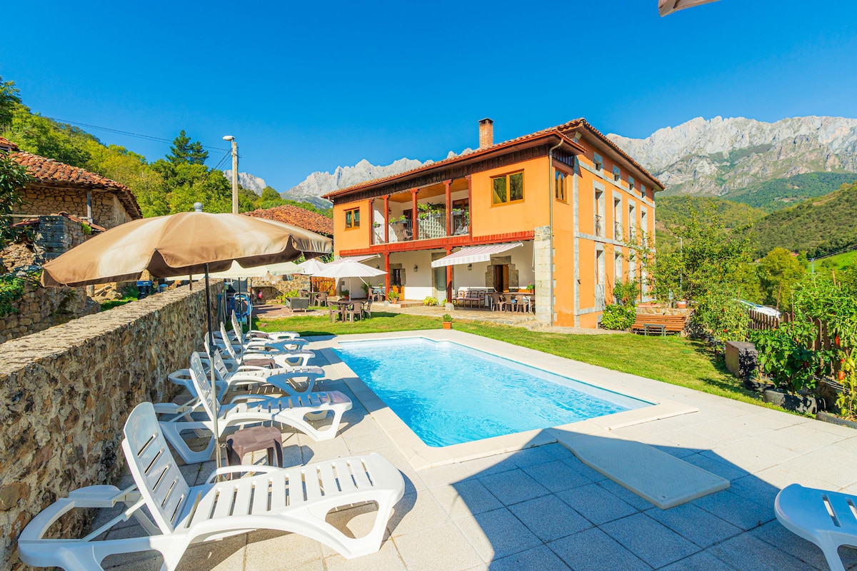 A spacious outdoor area features an inviting swimming pool surrounded by sun loungers and umbrellas. The orange-colored building, with large windows, overlooks a scenic landscape of mountains and greenery, providing a serene backdrop for relaxation and leisure activities.