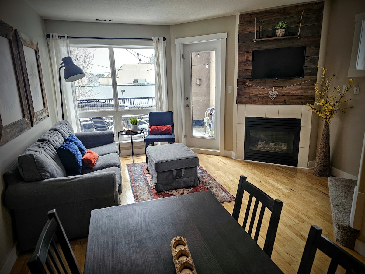 A comfortable living area is presented with a gray sofa, an ottoman, and a dining table. Natural light enters through large windows, illuminating the wooden flooring. A modern fireplace and a mounted TV are visible, enhancing the cozy atmosphere. A door opens to the patio.