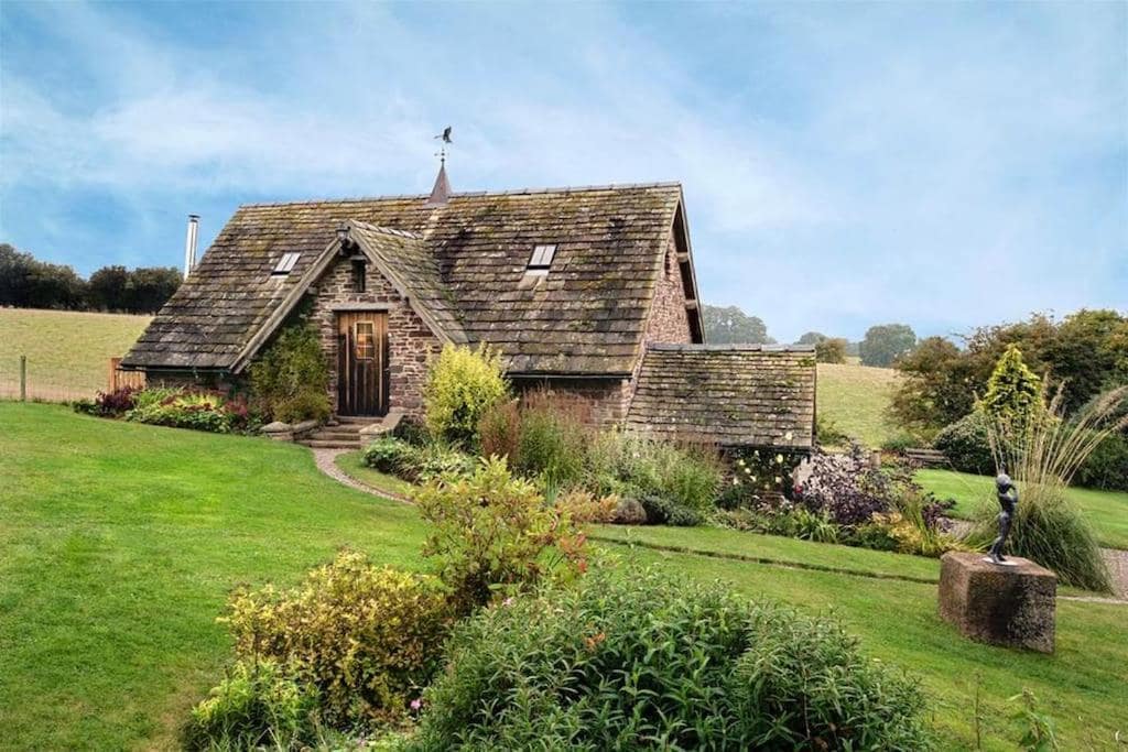 A traditional timber-framed cottage sits amidst lush greenery and colorful flowerbeds. The sloped roof features wooden shingles, and a decorative weather vane is visible at the peak. A winding pathway leads to the front door, surrounded by shrubs and plants, enhancing the tranquil countryside setting.
