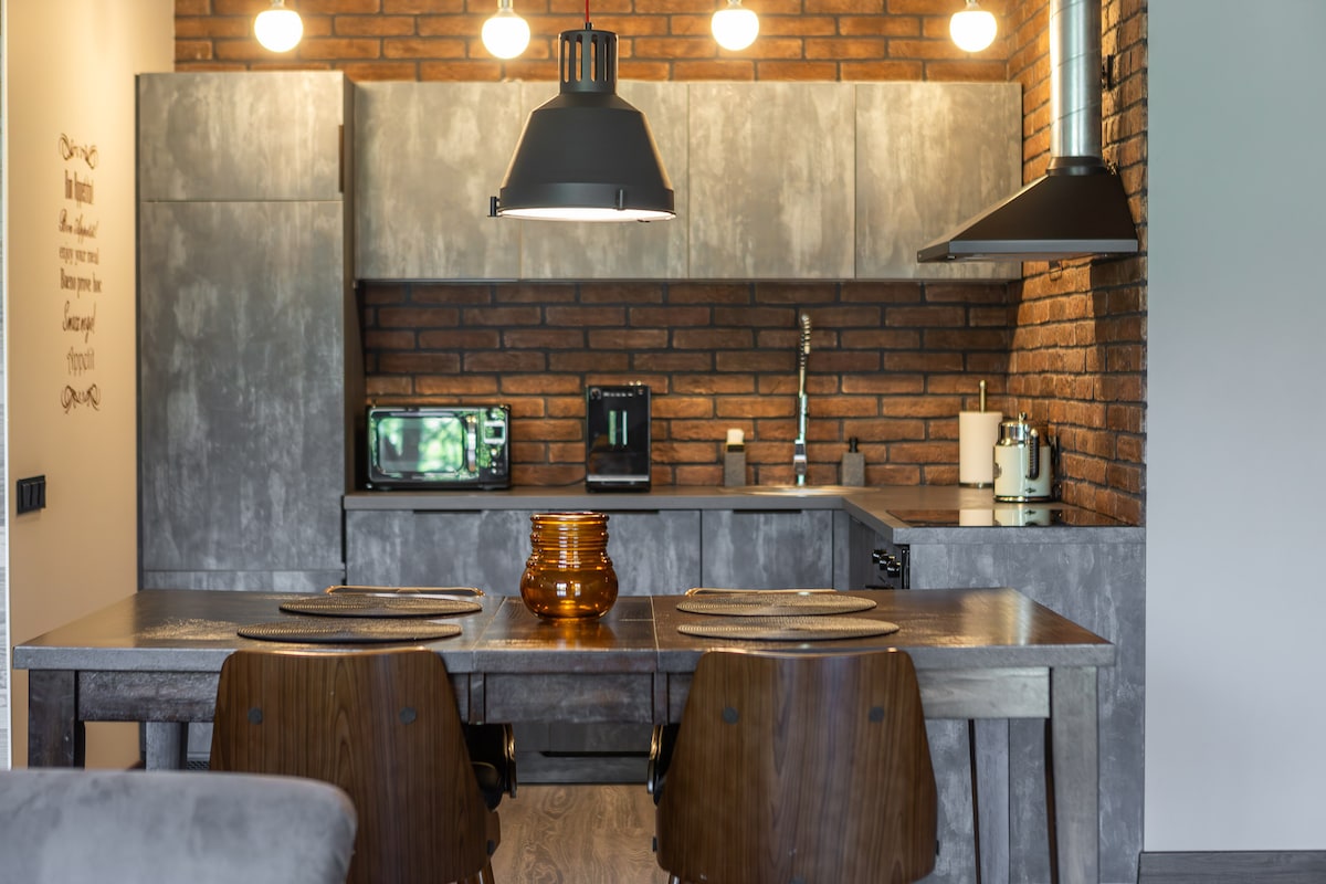 A modern kitchen is featured with industrial-style cabinetry in gray tones. A wooden dining table is set for two, accompanied by dark, stylish chairs. Warm lighting from pendant fixtures complements the exposed brick wall, adding character to the space.