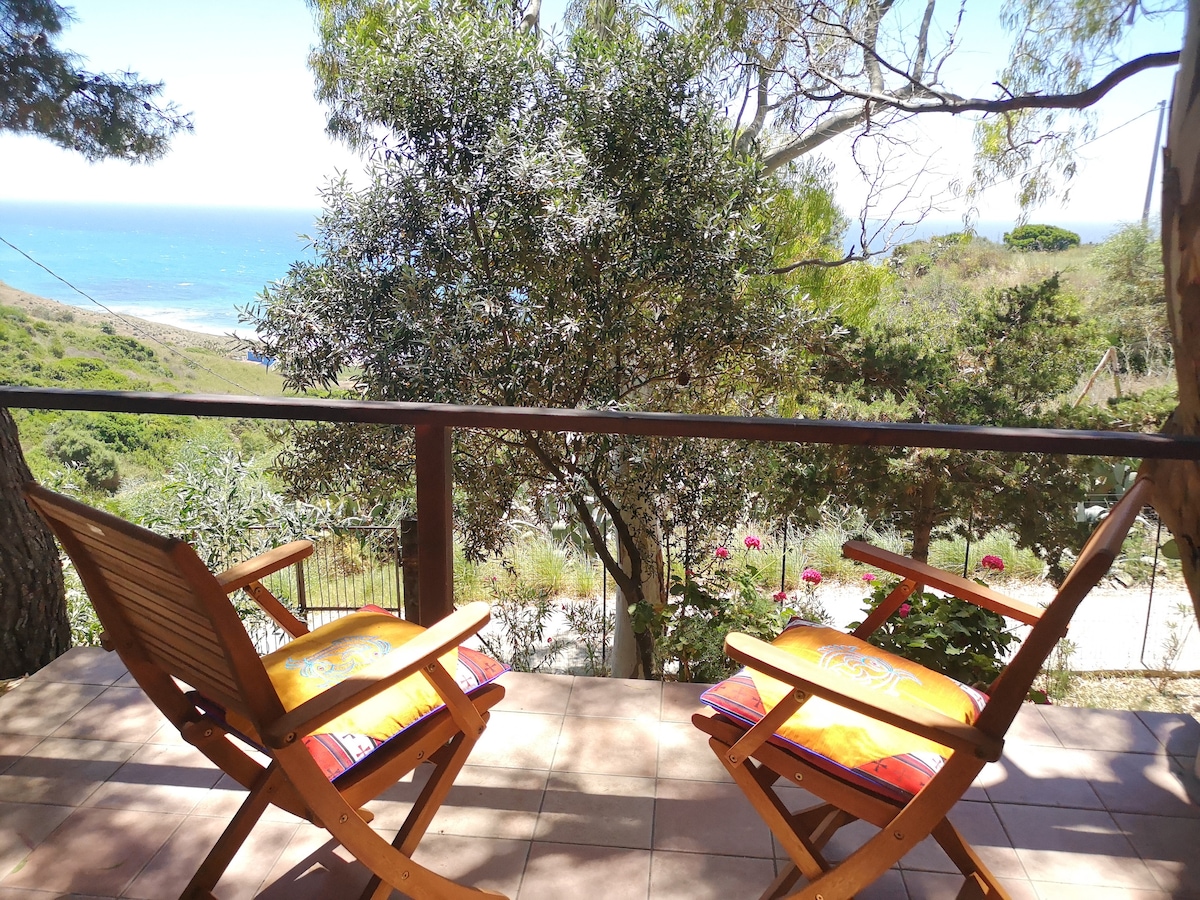 The outdoor terrace features two wooden chairs with bright cushions, overlooking a panoramic view of the sea and surrounding greenery. The space is surrounded by trees, providing a sense of tranquility and connection to nature.