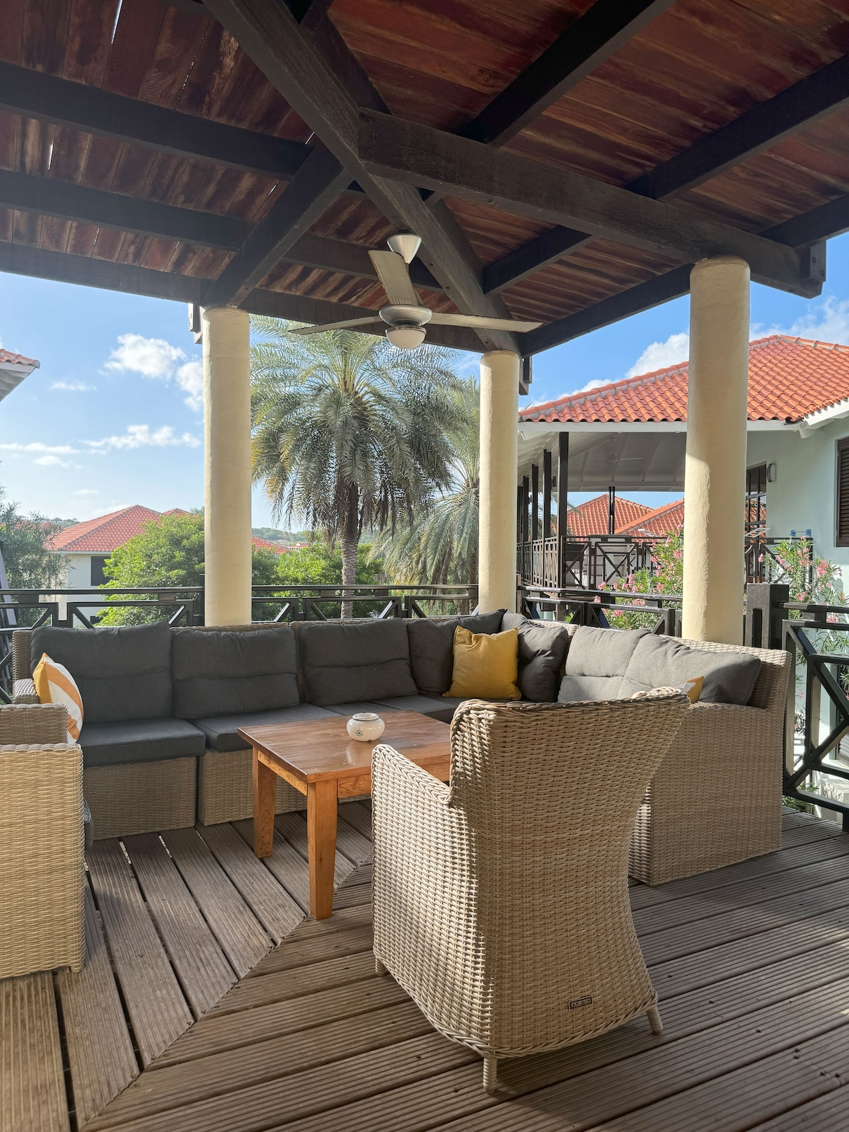 A spacious porch is furnished with comfortable seating, including a large sectional couch and a wicker chair. A wooden table sits in the center, with sunlight filtering through the overhead beams. Lush greenery and rooftops of nearby villas can be seen in the background.