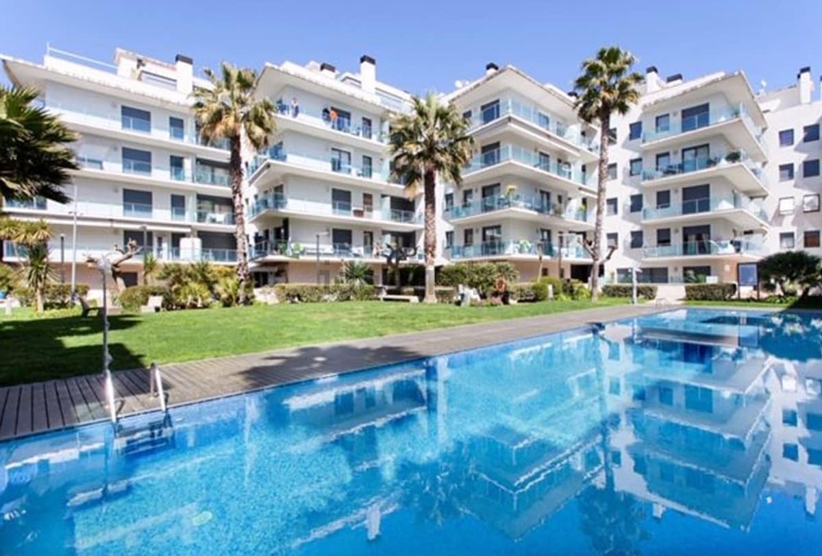 A residential complex is showcased with multiple white buildings surrounding a clear swimming pool. Lush palm trees and manicured lawns are visible in the foreground, reflecting the vibrant blue sky and serene atmosphere, creating an inviting outdoor space for relaxation and leisure.