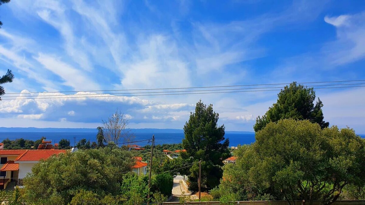 A panoramic view of the coastline is presented, with the sea stretching into the distance under a wide sky adorned with clouds. Lush green trees frame the foreground, while rooftops of nearby homes are visible, contributing to a serene natural environment.