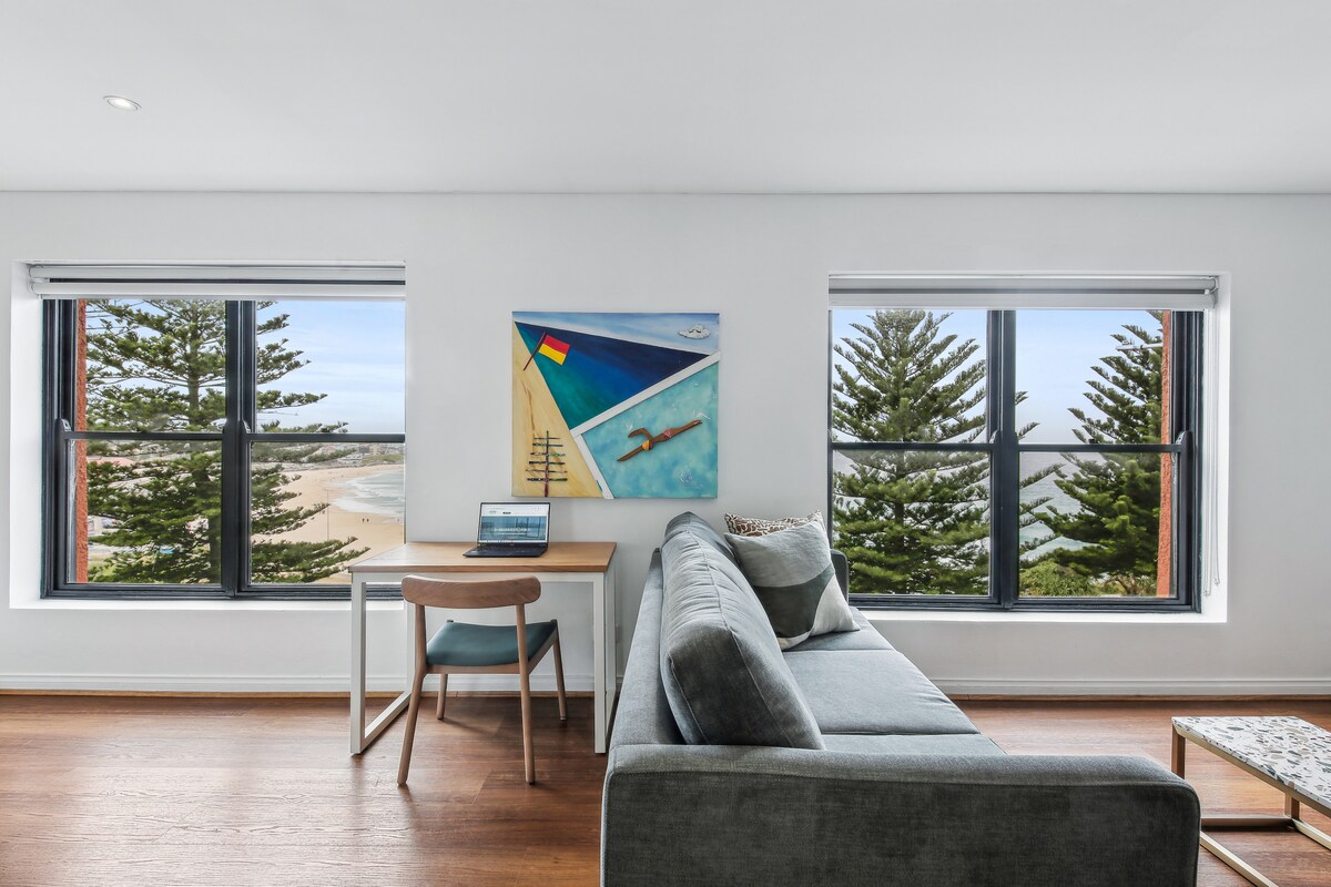 This living area features a light, modern design with a comfortable grey couch and a wooden table. Two windows frame the view of sandy beaches and greenery outside, allowing natural light to fill the space. A piece of artwork adds a colorful accent to the wall.
