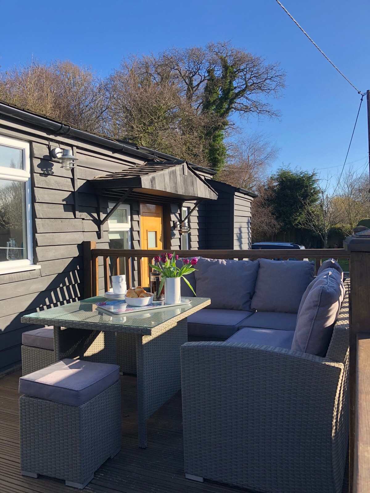 An outdoor decked area features a comfortable seating arrangement with a glass-top table and plush cushions. Soft pink flowers are arranged in a vase atop the table, creating a relaxed space for dining or socializing, surrounded by lush greenery under a clear blue sky.