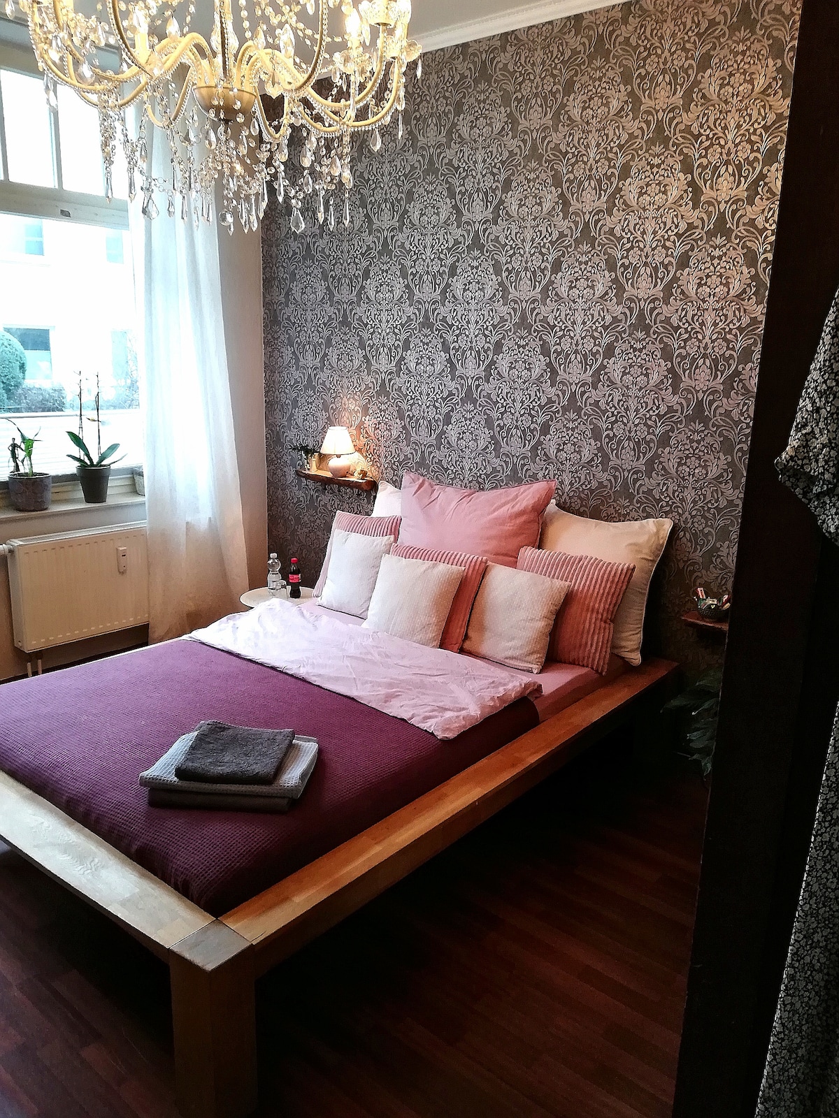 A cozy sleeping area features a wooden bed with pillows in various textures and tones. Elegant wallpaper with a floral pattern complements the room. Natural light enters through a window with soft curtains, and plants are positioned near the window, adding a touch of greenery.