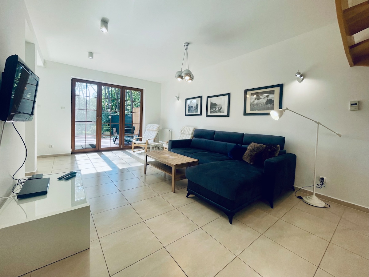 A spacious living area is arranged with a dark sectional sofa and a light wooden coffee table. Large glass doors provide access to the outdoor space, allowing natural light to illuminate the room. A TV is mounted on the wall, complemented by stylish wall art.