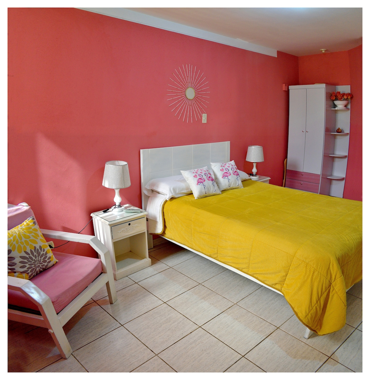 A spacious bedroom features a bright yellow bedspread on a large bed, complemented by decorative pillows. Pink walls are accented by a wall-mounted sunburst design. A bedside cabinet with a lamp and a cozy chair add to the room's comfort, while a wardrobe provides additional storage.