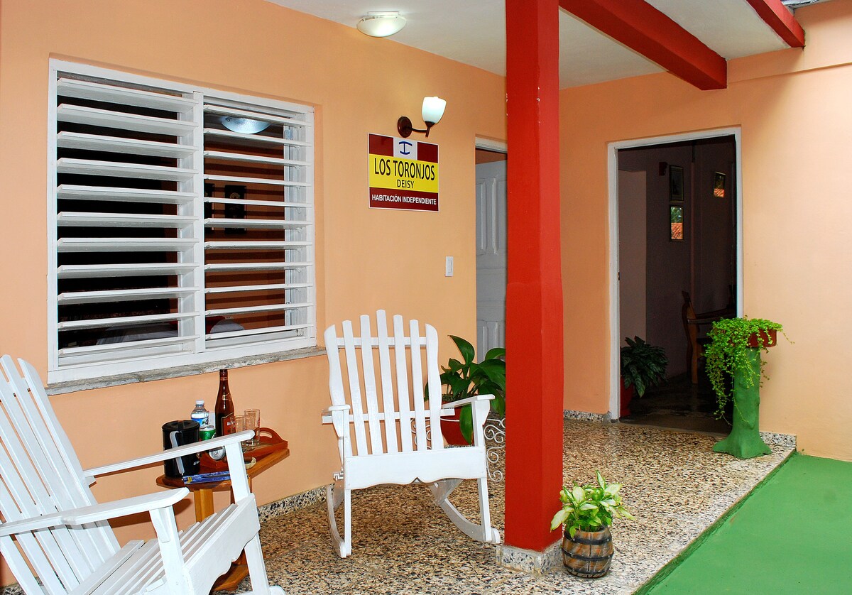 A shaded porch features two white rocking chairs positioned around a small wooden table. A sign displaying the property name is mounted on the wall above. Potted plants add touches of greenery, while a doorway to the interior is visible in the background.