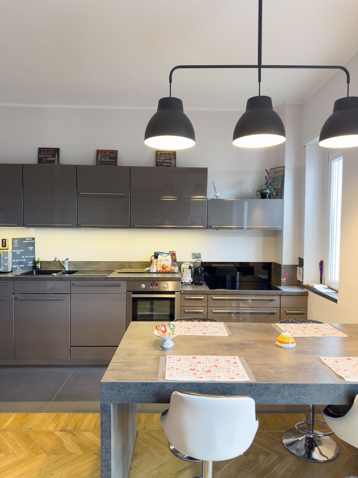 The kitchen features sleek, modern cabinetry in dark tones, complemented by a broad countertop. A dining table with white chairs is set with placemats, while overhead pendant lights provide ample illumination. The space is equipped with essential appliances, creating a functional cooking environment.