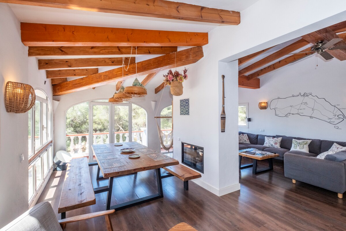 A spacious living area is highlighted by wooden beams and large glass doors, which allow natural light to fill the room. A long wooden dining table is flanked by benches, while a cozy seating arrangement features a sectional sofa facing the fireplace.