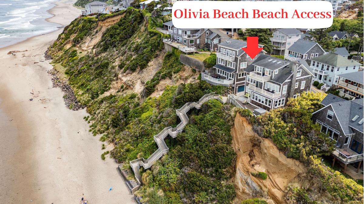 An aerial view displays the oceanfront home set against a backdrop of lush greenery and sandy beach. A winding staircase is visible, leading down to the beach access. The coastline curves gently, showcasing the expansive sandy shore and rolling waves.