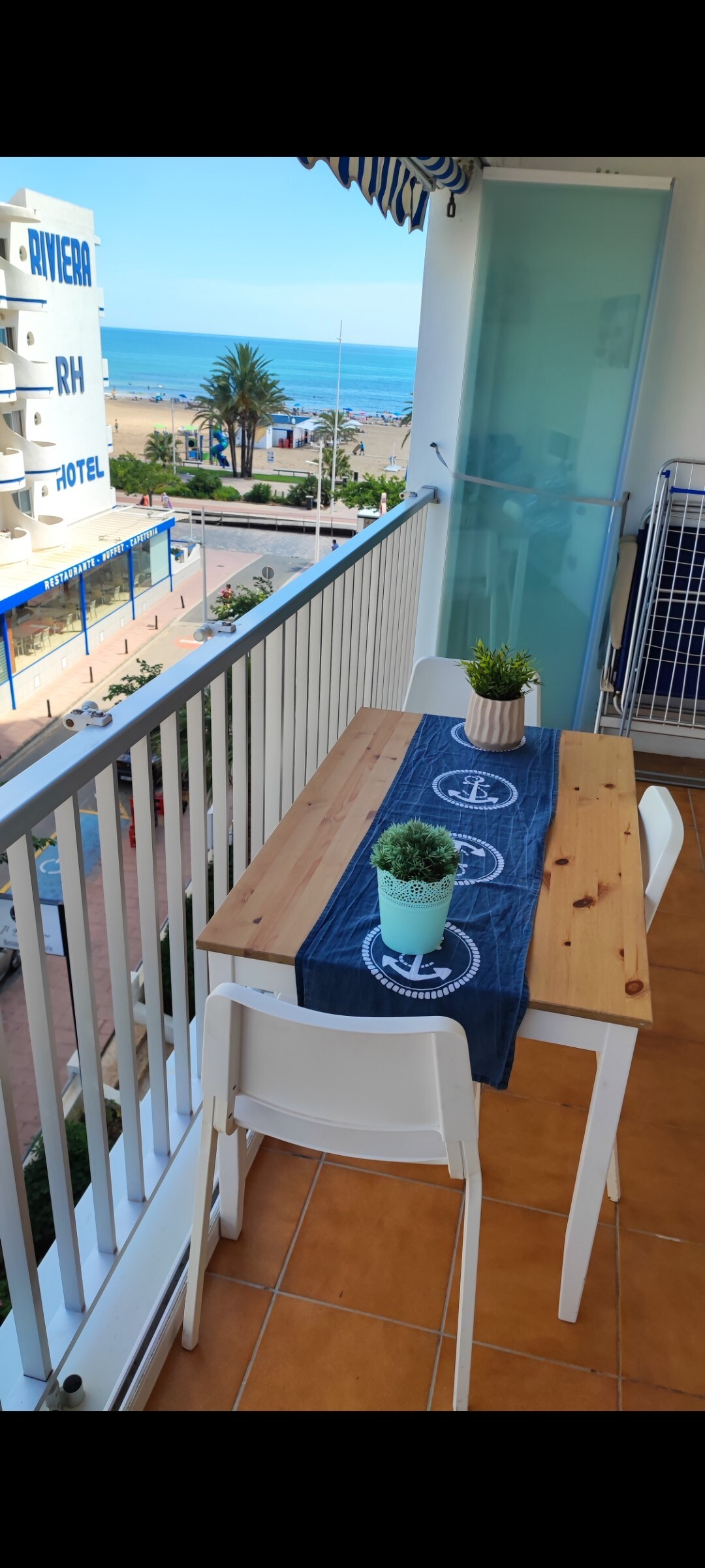 A small balcony is presented with a wooden table topped by two potted plants. Two white chairs are placed around the table, with the ocean visible in the background. Bright sunlight highlights the tiled floor, enhancing the outdoor dining space.