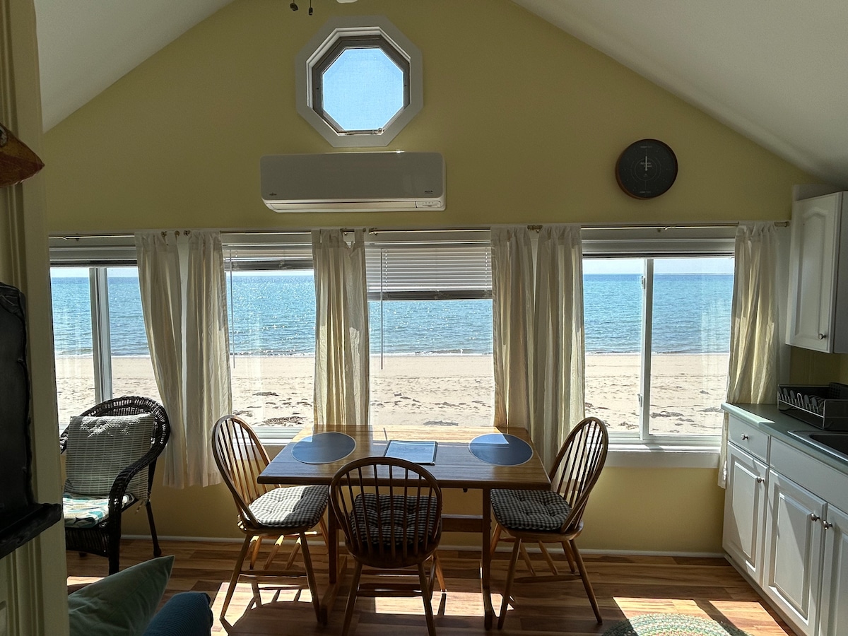 A light-filled dining area features a wooden table accompanied by four wicker chairs, situated near large windows that provide a view of the sandy beach and water. Sheer curtains gently frame the windows, enhancing the bright and airy ambiance.