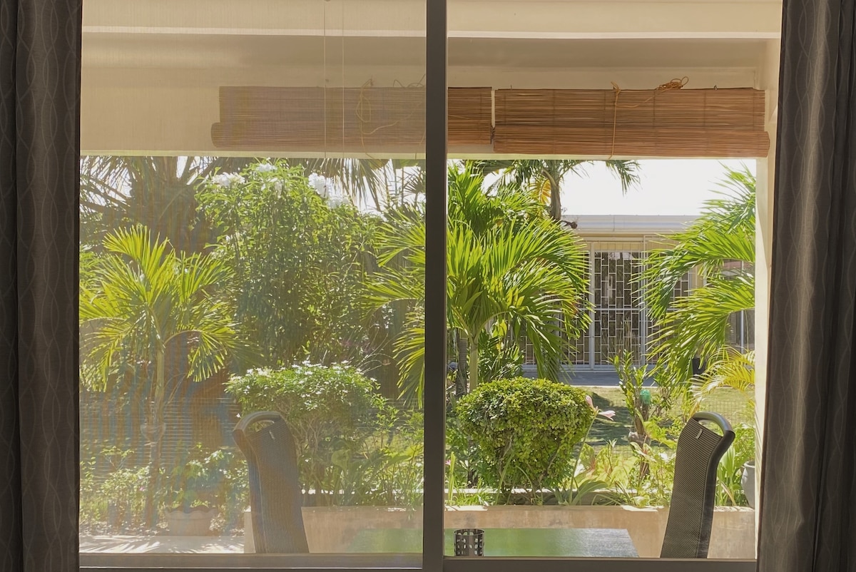 A large window offers a view of lush greenery, including palm trees and manicured bushes. Outdoor furniture is visible on a private terrace, inviting relaxation amidst a serene, tropical setting. Natural light filters through sheer window coverings, enhancing the indoor space's brightness.