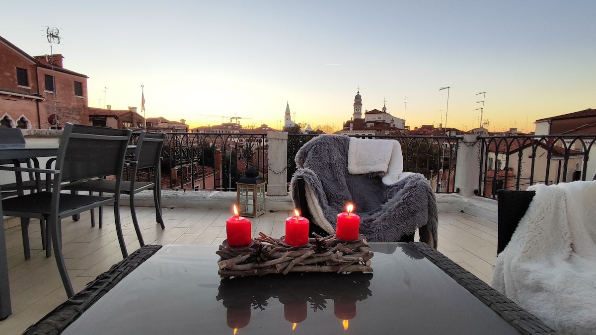 An outdoor terrace features a table with a decorative arrangement of candles. A cozy chair is draped with a soft blanket, surrounded by a scenic view of Venetian rooftops and a sunset illuminated sky.