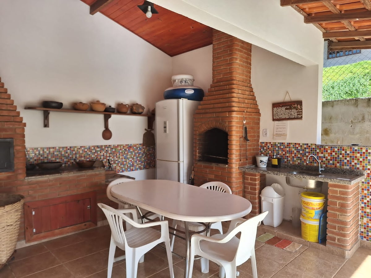 An outdoor kitchen area is featured, showcasing a stone grill and a large countertop made of stone. A round table is surrounded by white plastic chairs. Colorful tile accents decorate the walls, complemented by a traditional clay pot collection and a refrigerator against the back wall.