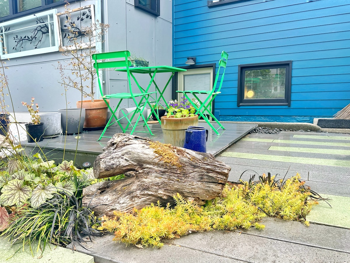 A small outdoor seating area is displayed, featuring two green bistro chairs and a matching table, inviting relaxation. Nearby, a rustic log adds natural texture, surrounded by colorful plants in pots and various ground cover plants, complemented by sleek decking.
