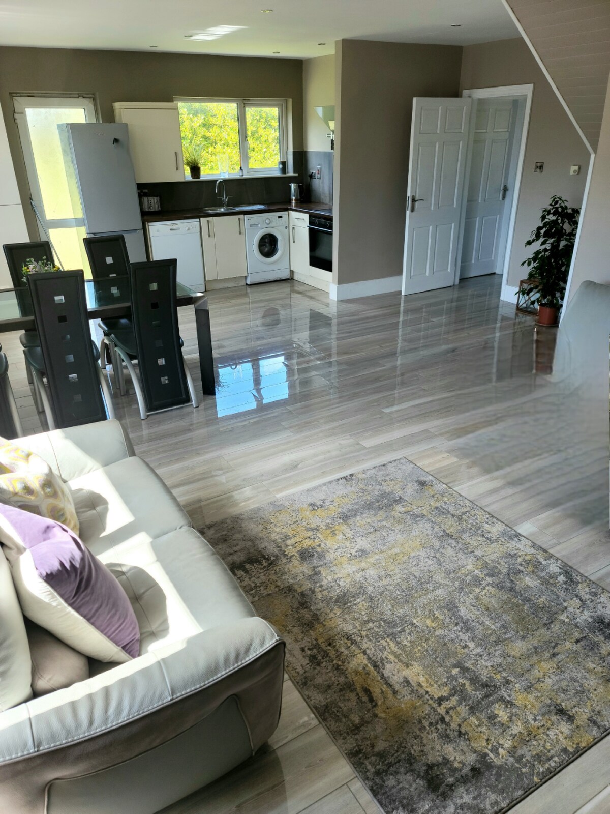 An open-plan living area is highlighted by polished floor tiles, reflecting natural light. A modern kitchen is visible in the background with white cabinetry and stainless steel appliances. A light gray sofa with colorful cushions is positioned towards a small area rug, enhancing the inviting layout.