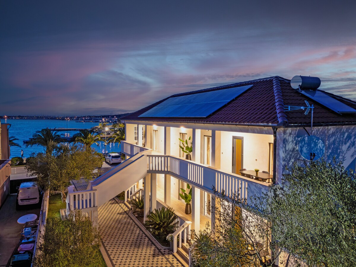 The modern building features a spacious balcony overlooking the sea, illuminated by soft evening light. Solar panels adorn the roof, while lush greenery surrounds the entrance. The tranquil water glimmers in the background, enhancing the serene coastal ambiance.