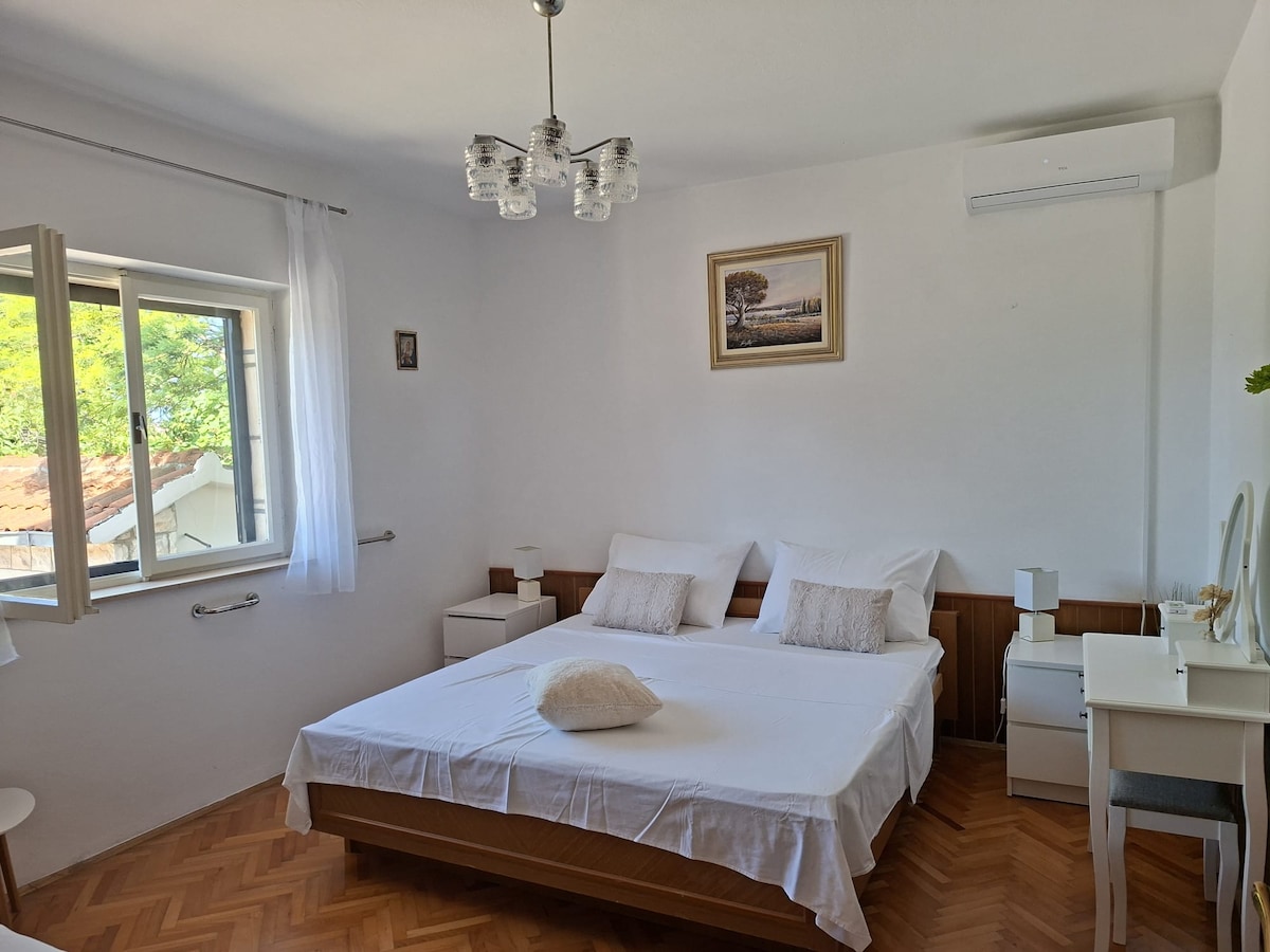 A bright bedroom is presented, featuring a spacious bed with neutral bedding. Two nightstands with lamps are positioned on either side, along with a vanity table. A window allows natural light to fill the room, while an air conditioning unit is mounted on the wall.