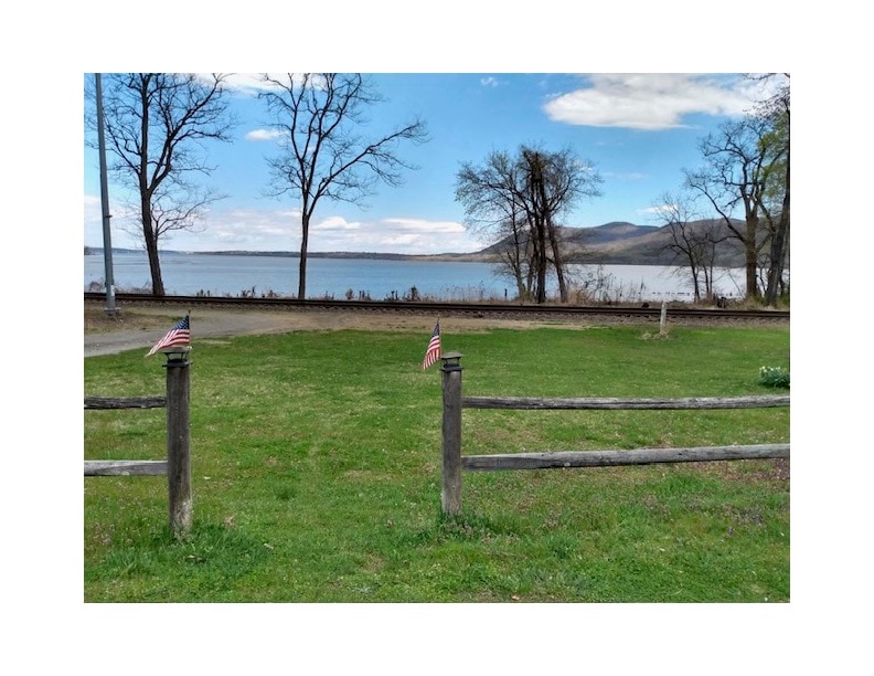 A scenic view of the Hudson River showcases lush green grass in the foreground. Wooden posts with small American flags mark the entrance, while tree branches gently sway in the breeze. The tranquil river stretches across the background, with distant hills creating a peaceful atmosphere.