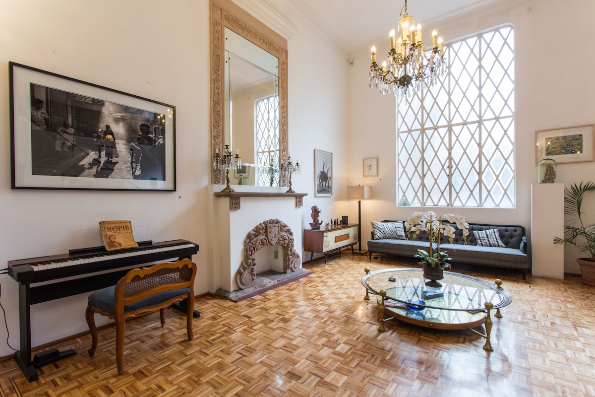 A spacious lounge area is highlighted by a grand chandelier and large, diamond-patterned windows allowing natural light to fill the room. A decorative mirror and artwork adorn the walls, while a piano and elegant coffee table add to the inviting atmosphere.