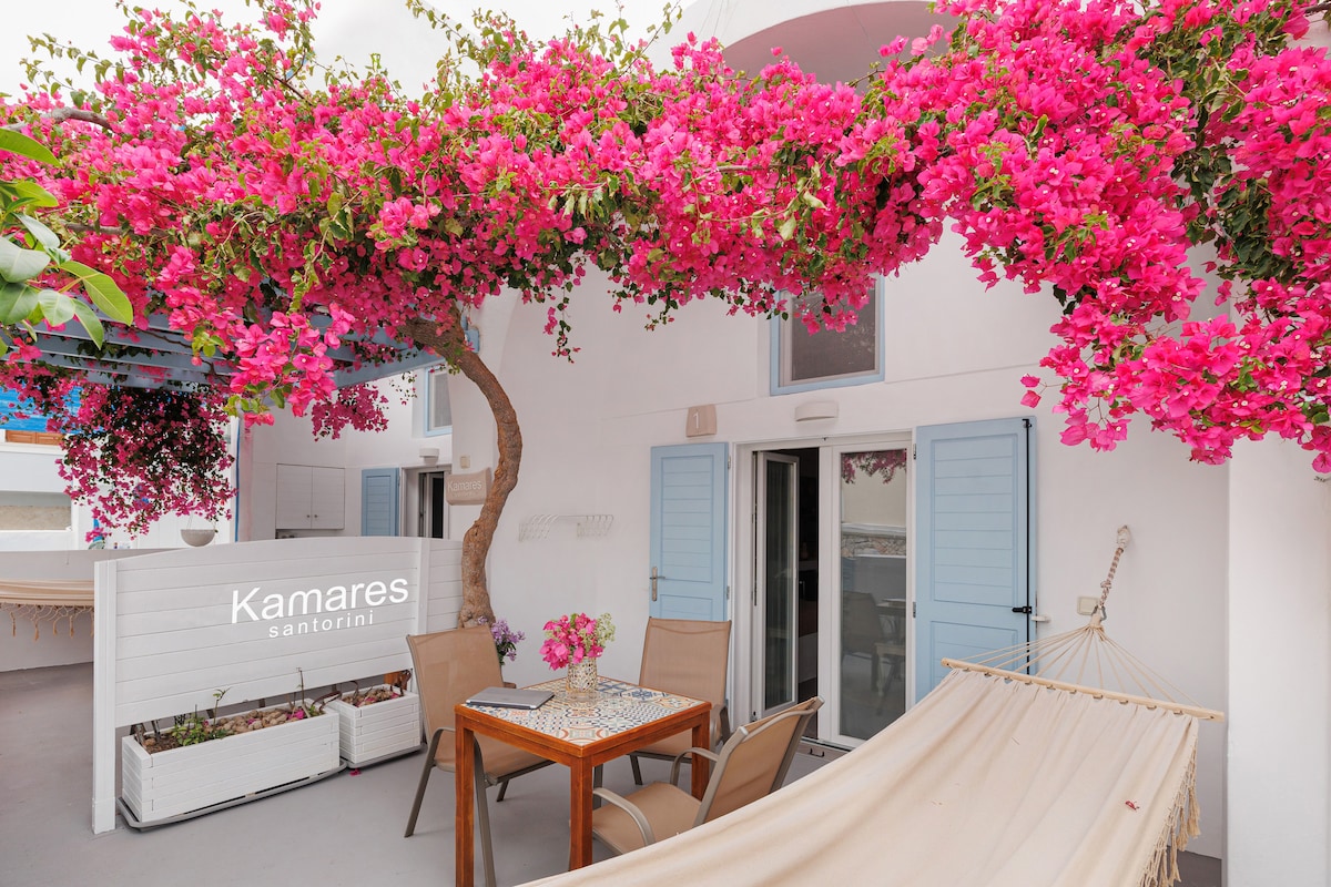 An outdoor terrace is adorned with vibrant pink bougainvillea cascading down, providing natural shade. Two chairs and a table are positioned on a light-colored floor, creating a cozy seating area. A hammock is visible, inviting relaxation in a tranquil environment.