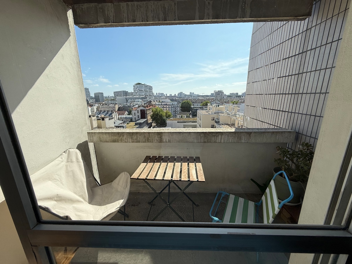 A private balcony features a rustic wooden table and two seating options, encouraging relaxation. A view of the urban skyline stretches into the distance, framed by a clear blue sky. The space is enhanced by greenery, creating a refreshing outdoor retreat.