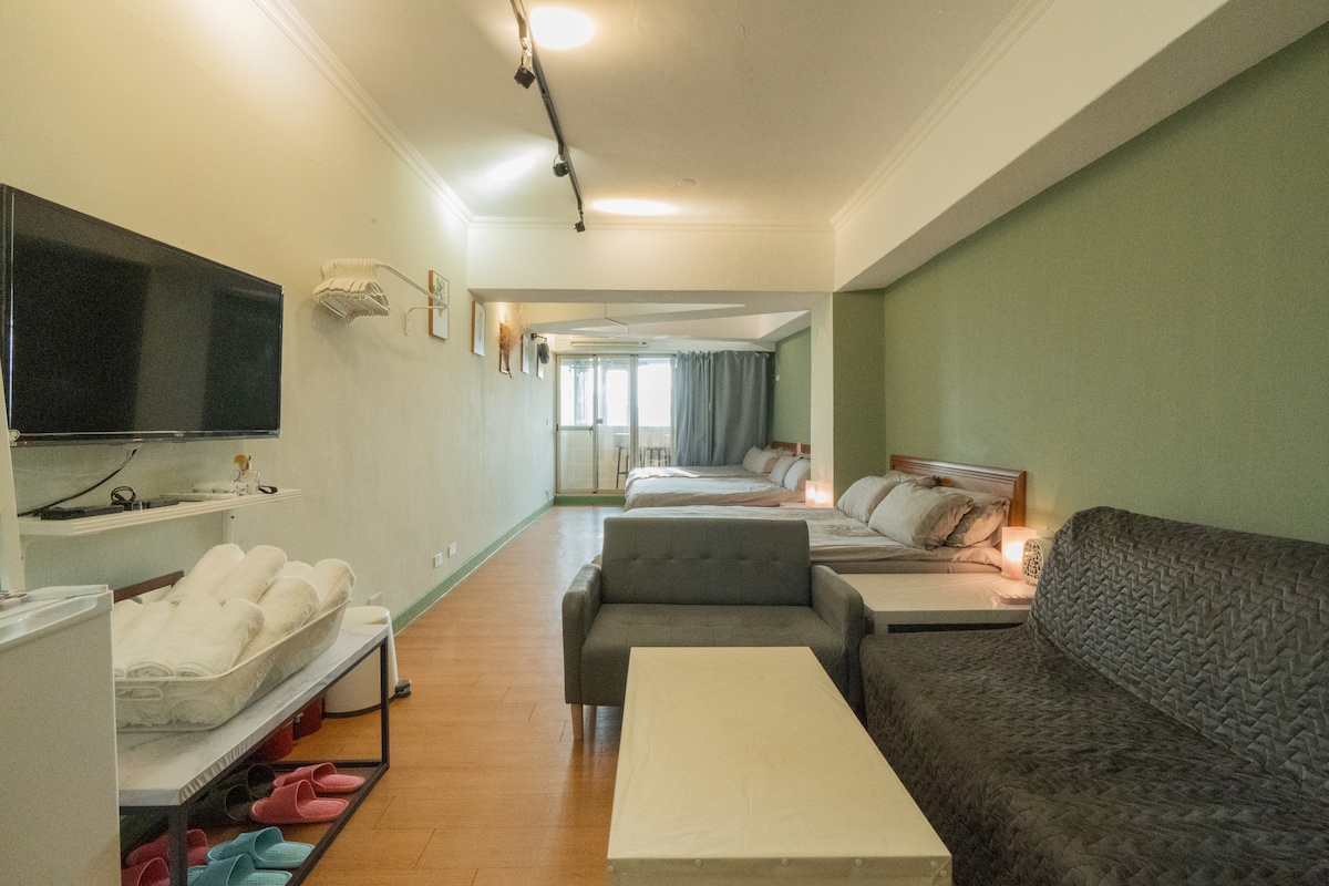 A spacious studio features distinct sleeping areas with comfortable beds surrounded by light green walls. A gray sofa and a coffee table are positioned in the living area, while a television and small refrigerator are visible along one wall.