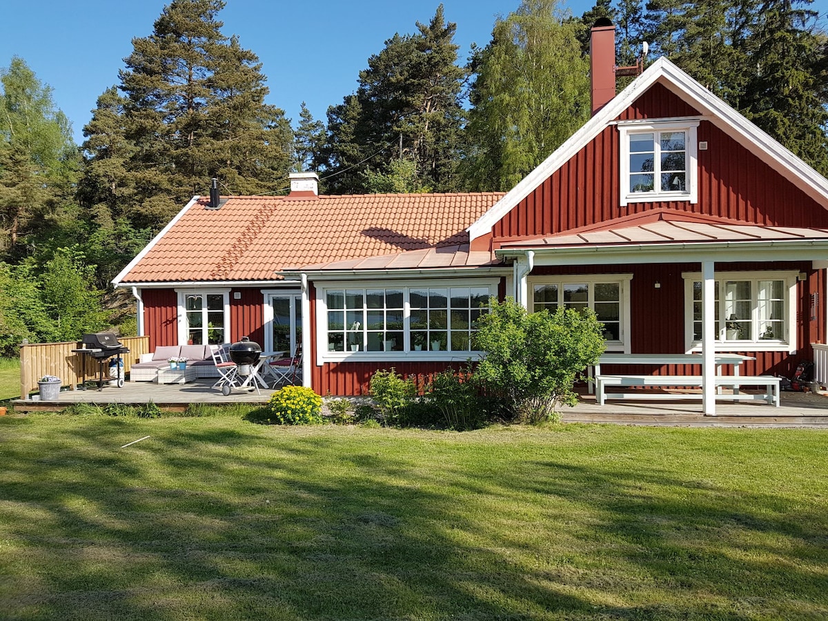 The classic Swedish-style house features a red exterior with white trim. Expansive windows provide ample natural light, and a covered porch offers seating. A garden area with well-maintained grass and a barbecue grill is visible, surrounded by trees for added privacy.