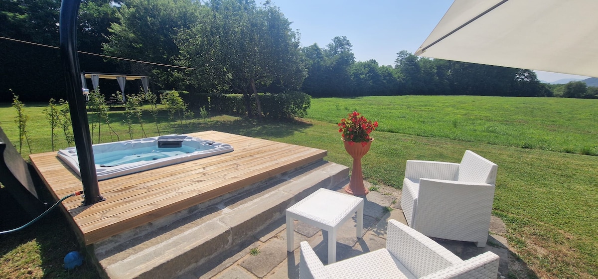 A wooden deck features a hot tub surrounded by lush greenery, providing a tranquil outdoor setting. Two white chairs and a small table are positioned nearby, with a potted flower adding a touch of color to the space. A sunshade offers comfort during sunny days.