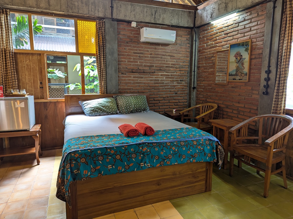 A cozy room features a spacious bed adorned with colorful bedding. Two wooden chairs are positioned beside the bed, and a small kitchenette is visible, complete with a refrigerator. Natural light enters through windows framed with patterned curtains, highlighting the exposed brick walls.