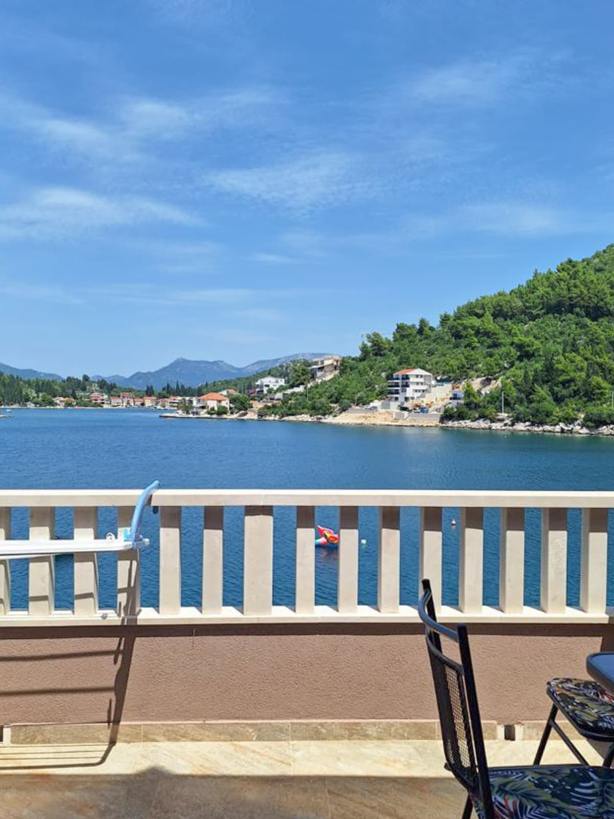A balcony view overlooks the serene waters, framed by green hills in the background. The scene captures a glimpse of the nearby village's buildings and a small boat gliding across the water. Sunlight bathes the area, creating a peaceful setting.