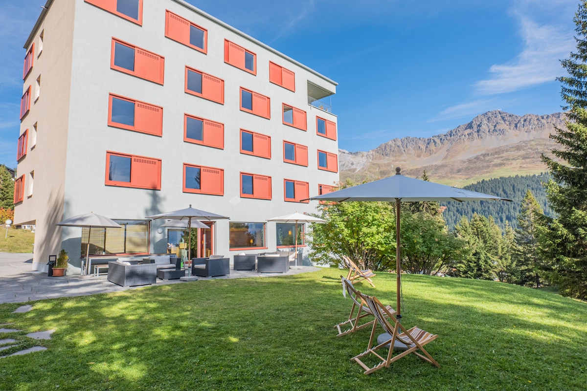 A multi-story building with modern architecture features large windows and bright red accents. In the foreground, a grassy area is adorned with lounge chairs and patio umbrellas, offering a relaxing outdoor space. The backdrop showcases beautiful mountain scenery under a clear blue sky.
