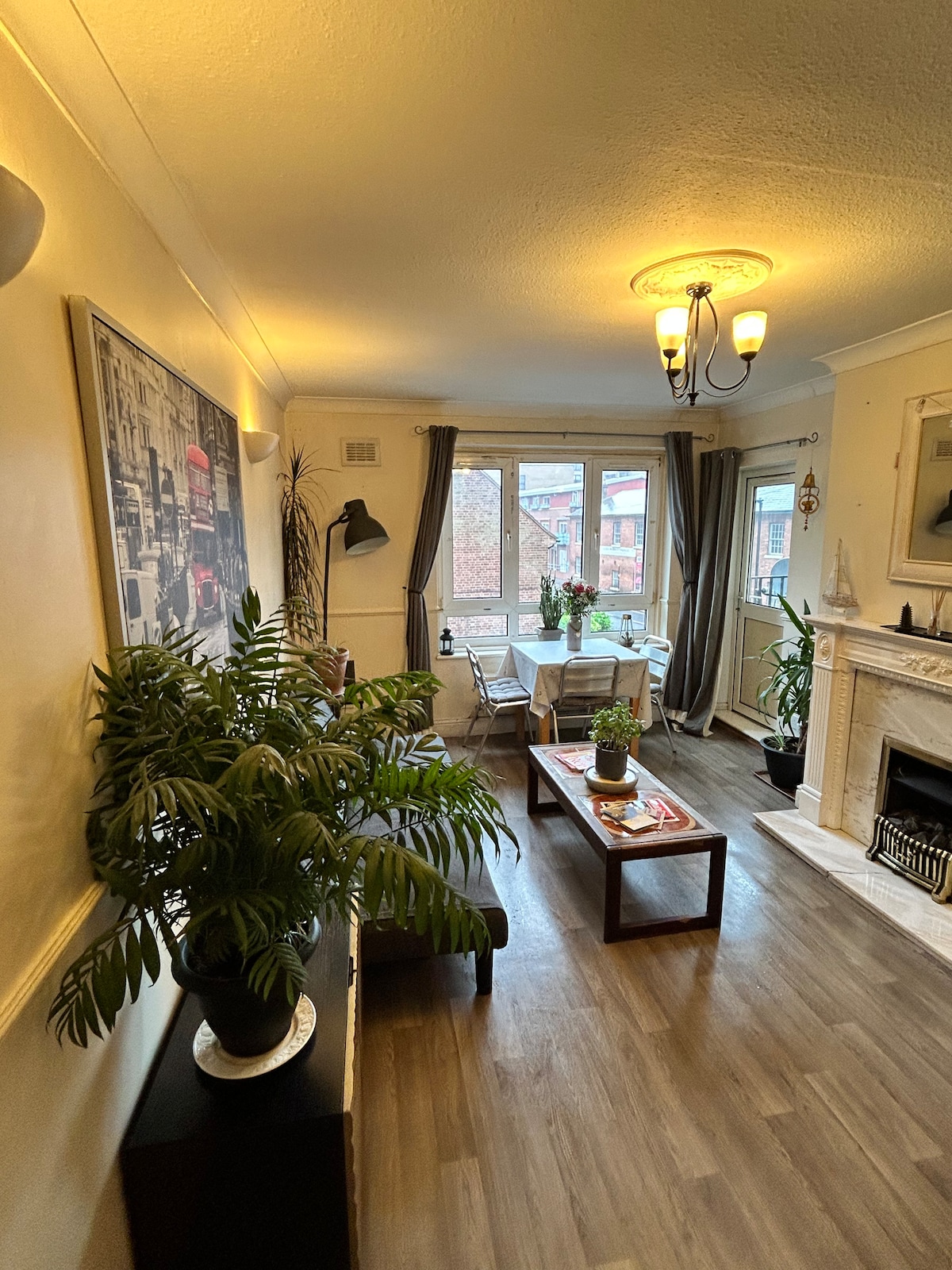 The living area features a stylish seating arrangement with a sofa and coffee table, complemented by decorative plants. A dining table is visible near large windows, allowing natural light to fill the space. A fireplace adds a cozy touch to the room's ambiance.