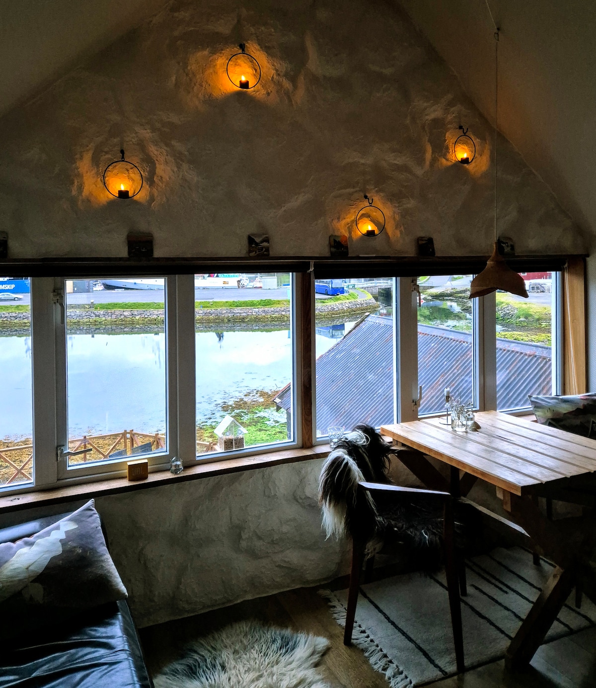A cozy nook features a large window with multiple panes, providing a view of the serene surroundings. Soft lighting is created by several wall-mounted fixtures, and a small wooden table is positioned near the window, accompanied by a sleek chair. Decor items add a touch of warmth.