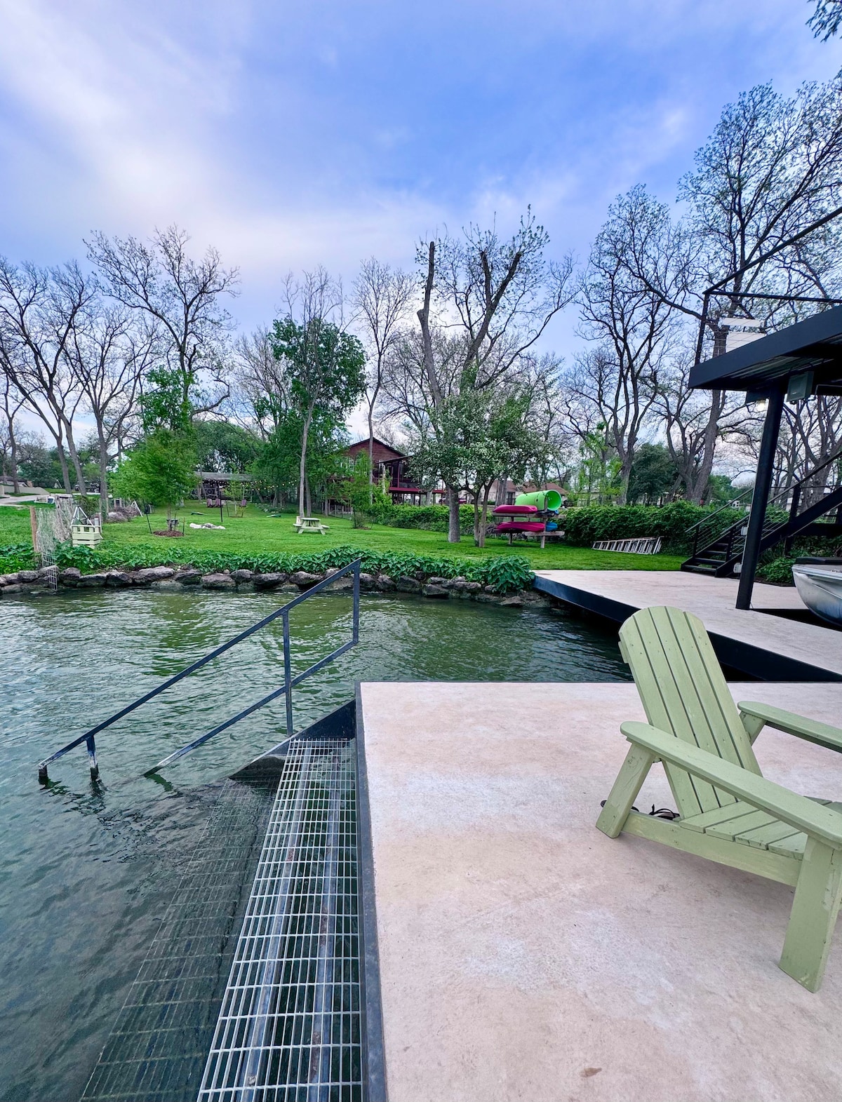 A serene waterfront view is presented, featuring green lawn areas dotted with trees in the background. A sturdy metal staircase leads into the water, with a wide deck visible, along with an adirondack chair positioned nearby, inviting relaxation by the lake.